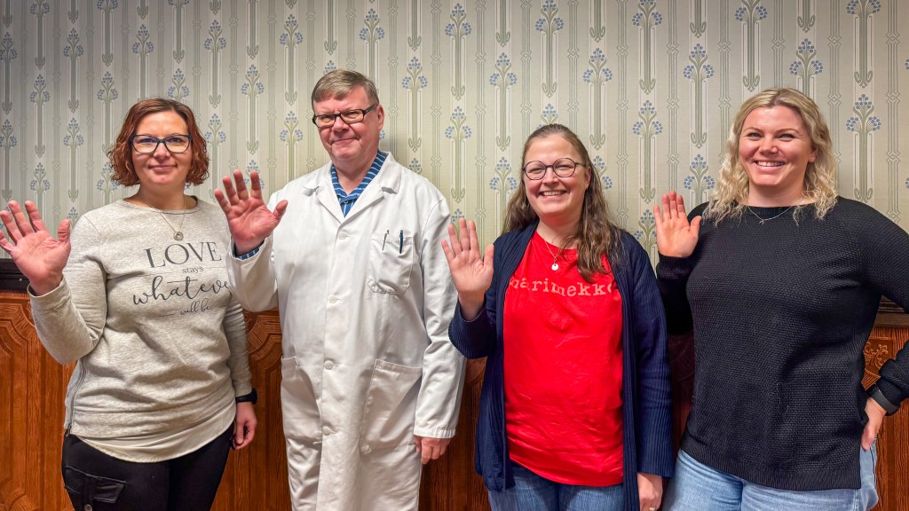 Neljä henkilöä seisoo rivissä, kaikki tervehtivät kätkemällä kätensä. He ovat sisätiloissa, seinät ovat vaaleat ja koristellut.